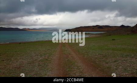 A dirt road leading towards a blue lake in North Mongolia under a cloudy sky Stock Photo