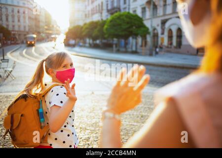 Life during covid-19 pandemic. elegant mother and child with masks and yellow backpack say goodbye before going to school outdoors. Stock Photo