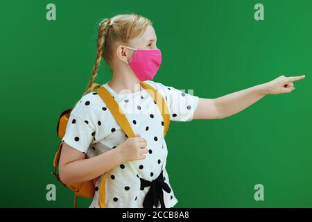 Life during coronavirus pandemic. modern pupil in white polka dot blouse with yellow backpack and pink mask pointing at copy space against chalkboard Stock Photo