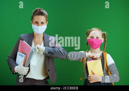 happy modern female teacher and pupil with masks and textbook greeted with elbow bump against chalkboard green background. Stock Photo