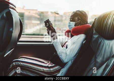 Side view of a young black woman with braids and in a virus protective mask sitting on the seat armchair of a regular bus and using her cellphone Stock Photo