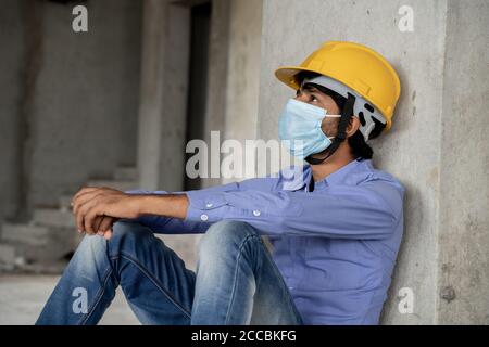 Head shot side view of construction worker seeing outside from construction building window in sad - Industrial worker in a hardhat with medical mask Stock Photo