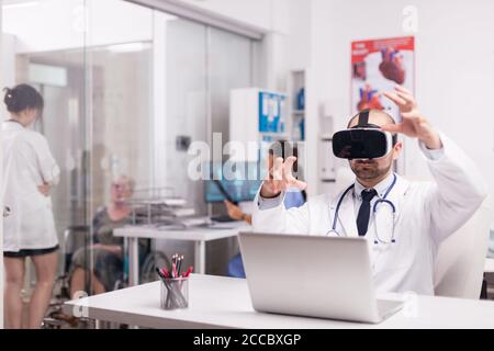 Young physician using virtual reality goggles in hospital office. Invalid senior woman in wheelchair talking with female doctor on clinic corridor. Nurse in blue uniform holding patient x-ray. Stock Photo