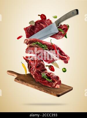 Flying pieces of raw steaks, with ingredients for cooking, served on woodenboard. Knife cuting the meat. Concept of food preparation in low gravity mo Stock Photo