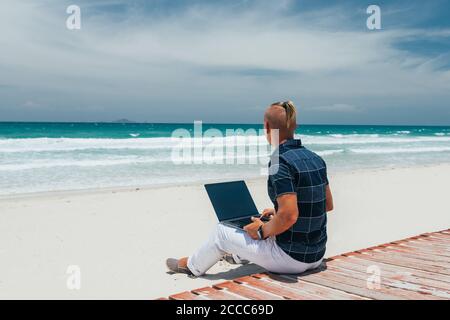 Young guy working using a laptop, sitting on the seashore on a sandy beach.Looking at the sea.  Businessman working on remote work. Blogger, freelance Stock Photo