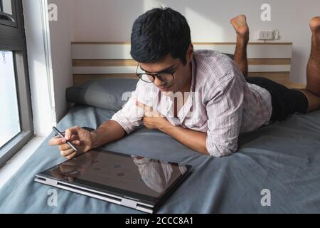 Young businessman with glasses lying on a bed and working with a graphic tablet Stock Photo