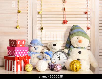 Snowmen, teddy bears and present boxes near alarm clock. Christmas decorations in festive room. Celebration and New Year decor concept. Toys placed on bureau on wooden wall and garlands background Stock Photo