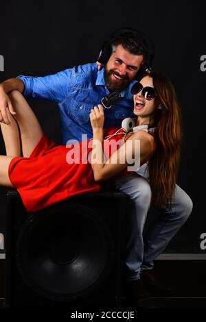 Partying and music concept. Music fans with happy faces enjoy music and sing. Couple in love wears headphones and holds microphone. Man with beard and girl sit on boombox on black background. Stock Photo