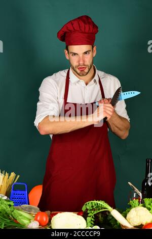Cuisine and cooking concept. Cook works in kitchen with vegetables and tools. Man in cook hat and apron sharpens blades. Chef with serious face holds blue and metal knives on dark green background. Stock Photo