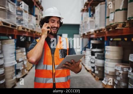 Successful factory manager tracking dispatch details using digital tablet while in conversation over smartphone in warehouse Stock Photo
