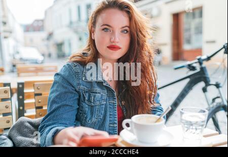 Portrait of sad red curled long hair caucasian woman sitting on a cozy cafe outdoor terrace on the street and looking at the street. Young woman takin Stock Photo