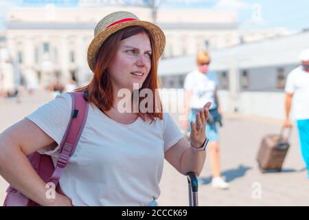 Irritated young red haired tourist woman in a hat with a backpack and suitcase stands at the train station Stock Photo