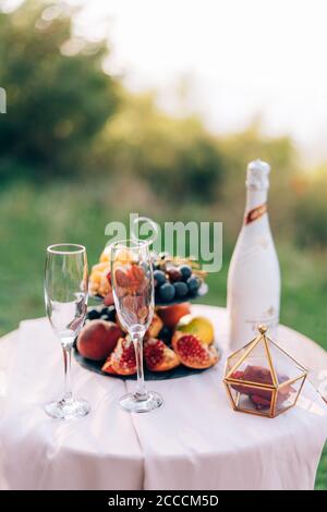 A table with a white tablecloth and two champagne glasses and a bookcase with fruits in nature. Stock Photo