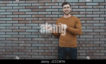 The horizontal look of an attractive young man who advertises something new, points his finger to the side, stands against a brick wall. People and li Stock Photo