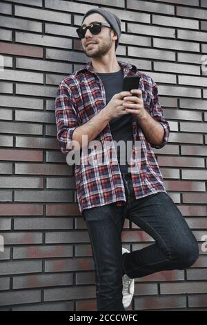 Hipster sms texting phone app in city street on brick wall background. Amazing man holding smartphone in smart casual wear standing. Stock Photo