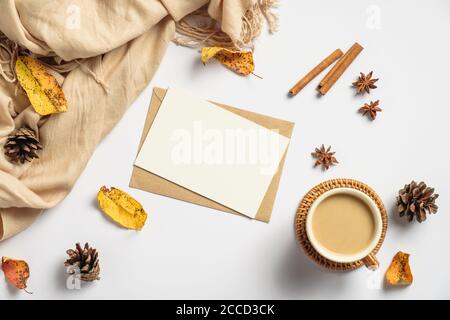 Autumn flat lay composition. Craft paper envelope, blank card mockup, coffee cup, cinnamon sticks, beige scarf, pine cones on white background. Autumn Stock Photo