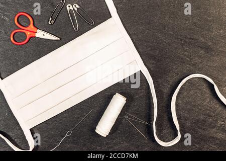 Homemade white cotton face mask with needle and thread on a dark background with view from above. Stock Photo