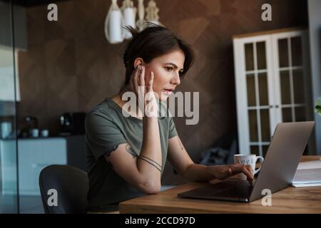 Attractive pensive young woman working on laptop computer while sitting at the table at home Stock Photo
