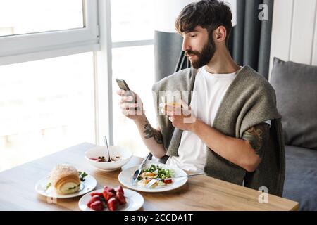 Attractive young bearded man using mobile phone while having healthy tasty dinner at home Stock Photo
