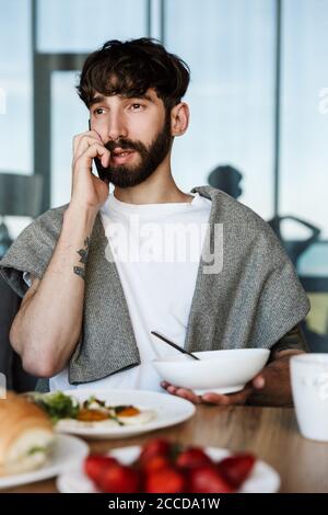 Attractive young bearded man using mobile phone while having healthy tasty dinner at home Stock Photo