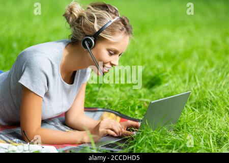 Young woman - watching educational webinar on laptop, lying on green grass in city park Stock Photo