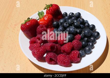 White plate with mixed berries including strawberries, raspberries and blueberries on a wooden table Stock Photo
