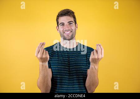 Young handsome man over isolated yellow background doing money gesture with hands, asking for salary payment, millionaire business Stock Photo