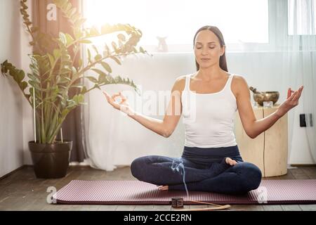 calm yogi female sit on the floor in lotus pose, meditate at home during quarantine, every morning begins from yoga, she use aroma sticks