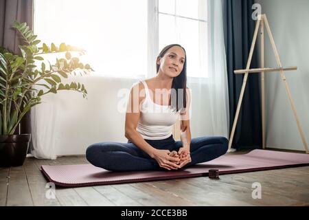 relaxed woman have rest after yoga exercises, sit on the floor with crossed legs, in sport clothes at home