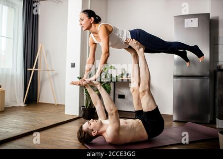 young caucasian married couple performing an acro yoga pose at home, man supporting the woman on his legs and arms, stretching and doing exercises tog Stock Photo