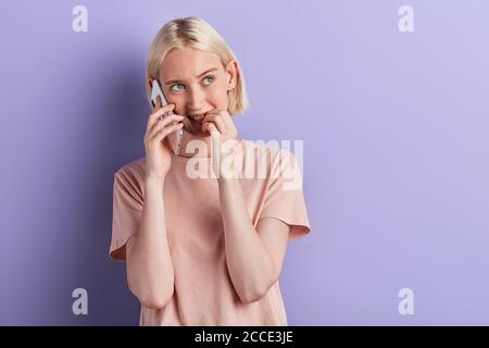 pensive woman talking on the smartphone, biting her nails, looking aside,thinking about questions , suggestion, offer, problem over violet isolated ba Stock Photo