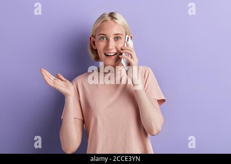 cheerful, emotional, astonished girl having smart phone cellphone in hand looking at camera gesturing with palm isolated on violet background. happine Stock Photo