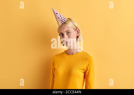 A young unhappy depressed woman in a birthday hat , stylish sweater, jumper looking at the camera. close up photo. isolated yellow background, studio Stock Photo