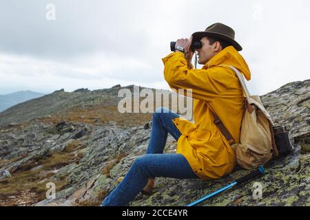 stylish hikers in raincoat, green hat with backpack holding binoculars sitting on top of the mountain and looks into the distance.close up side view p Stock Photo