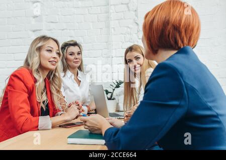 Team work process. Multiracial group of young women collaborating in open space office. Red-haired mentor coach speaking to young people, teaching aud Stock Photo