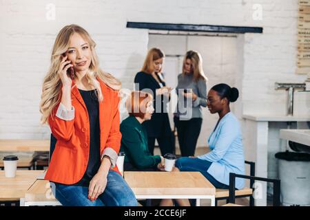 Attractive female journalist in red jacket looking at camera while chatting with friend on phone, while her girlfriends communicating on background Stock Photo