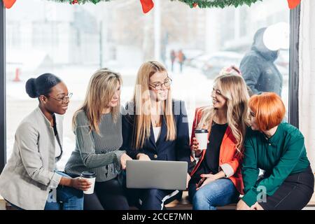Multiracial group of female students sitting together at a window sill with coffee and laptop for researching information for their project Stock Photo
