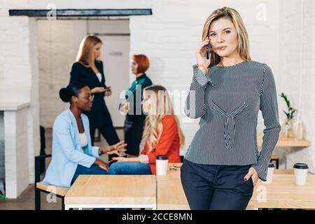 Caucasian mid-aged female manager, casual dressed, using smartphone in well-lit white office interior with her co-workers on background Stock Photo