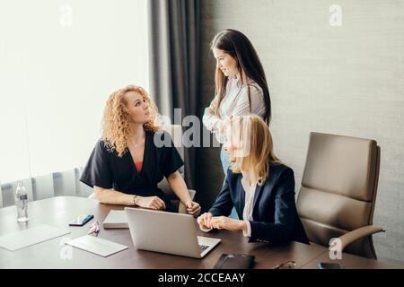 Female business team of young attractive employees discuss online project in modern office, collaborating using laptop and notepads for notes. Stock Photo