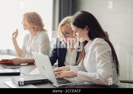 Three caucasian female students or interns in white shirts , intern using laptop, working on pc, studying in university, typing, browsing websites, ap Stock Photo