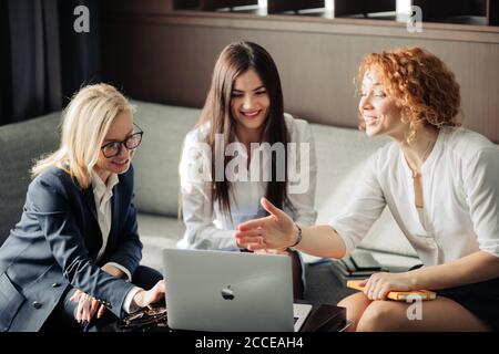 Three beautiful female fashion designers working as a team. Startup partners sitting at cafe, using laptop, discussing models of women wear, plans and Stock Photo