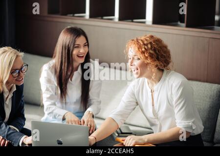 Red haired female corporate mentor helping two young women employees with computer work in open space office, close up. Stock Photo
