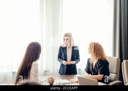 Elegantly dressed business women in office having conversation and using technology, focus on blonde woman telling something to her coworkers. Stock Photo
