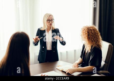 Elegantly dressed business women in office having conversation and using technology, focus on blonde woman telling something to her coworkers. Stock Photo