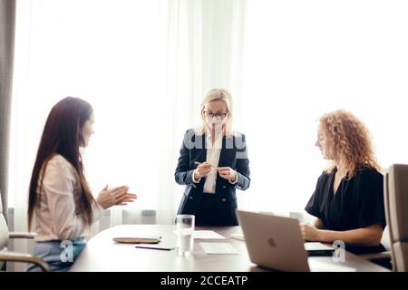 Female colleagues discussing work plans together, sitting at office table. Female red-haired intern asks questions to blonde head of department. Chief Stock Photo
