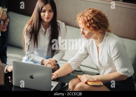 Three beautiful female fashion designers working as a team. Startup partners sitting at cafe, using laptop, discussing models of women wear, plans and Stock Photo