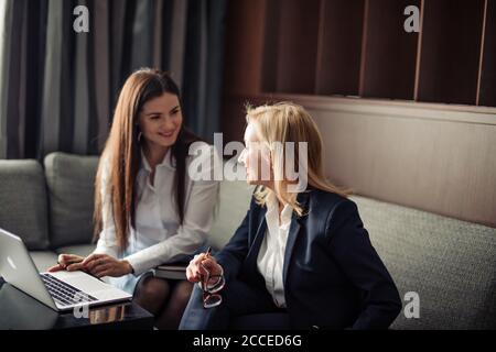 Two smiling female employees co-working on line with a laptop sitting on couch at office. Stock Photo