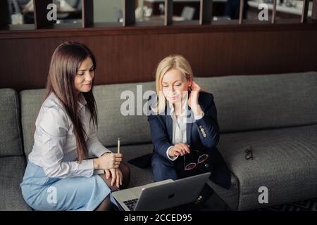 Blonde woman talking with her brunette business partner discussing data on laptop computer, two women entrepreneurs sitting on grey couch in business Stock Photo