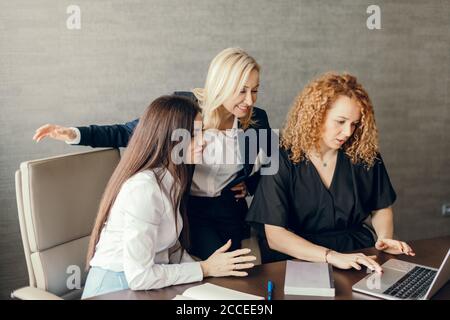 Group of three young female marketing company trainees share ideas with each other, looking at the laptop screen, while sitting in coworking space, cl Stock Photo