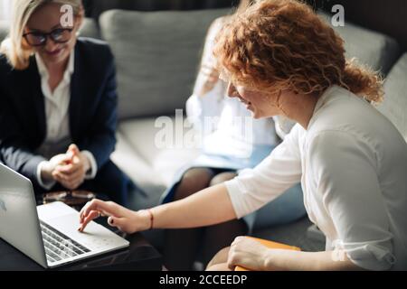 Group of three young female marketing company trainees share ideas with each other, looking at the laptop screen, while sitting in coworking space, cl Stock Photo